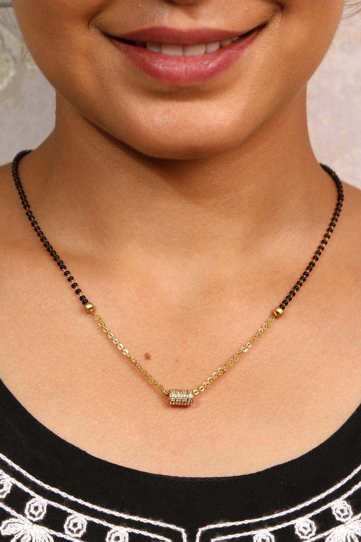A woman smiling, wearing a gold diamond-studded mangalsutra with black beads and a small cylindrical pendant.