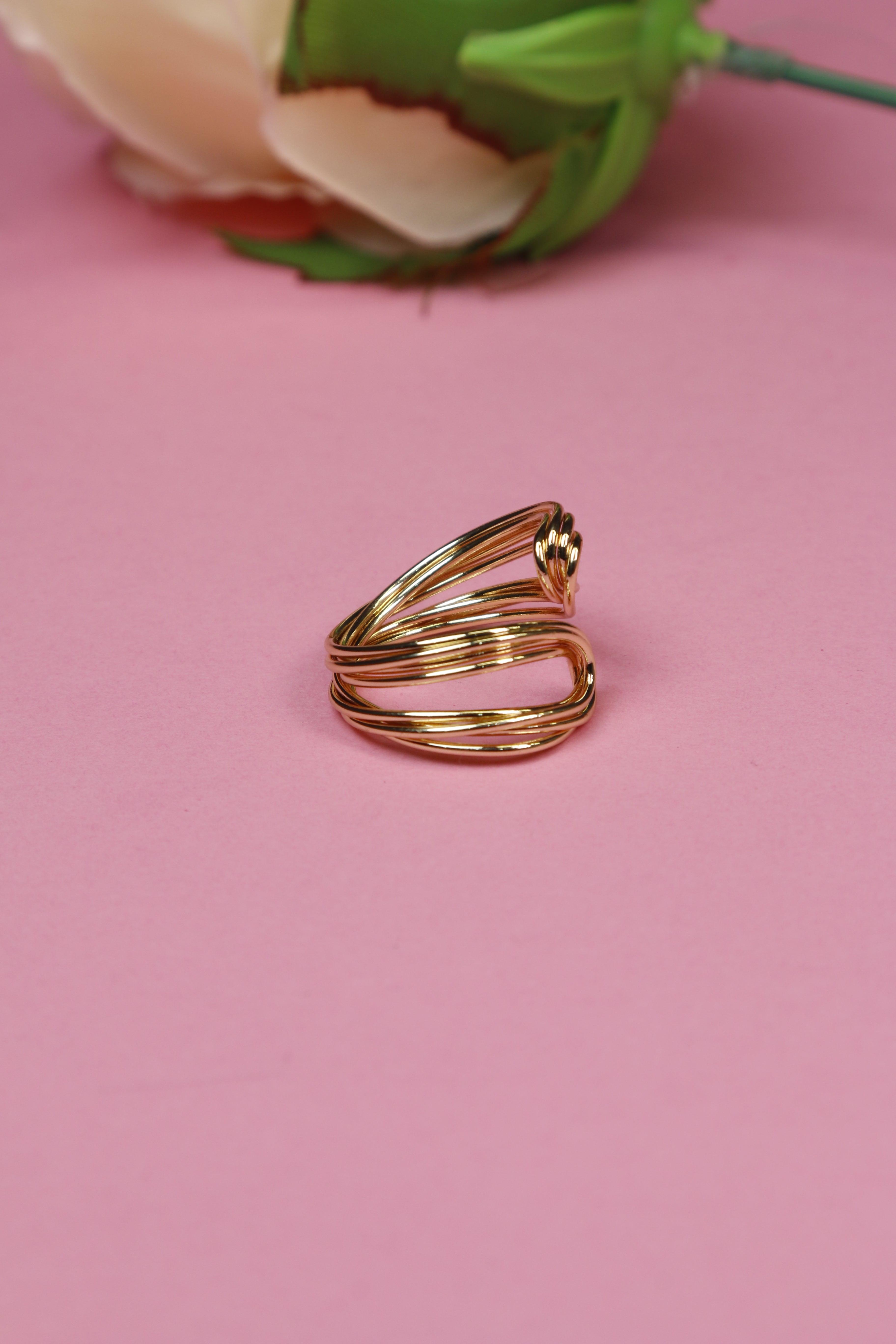Gold ring with flowing design, placed on a pink background.