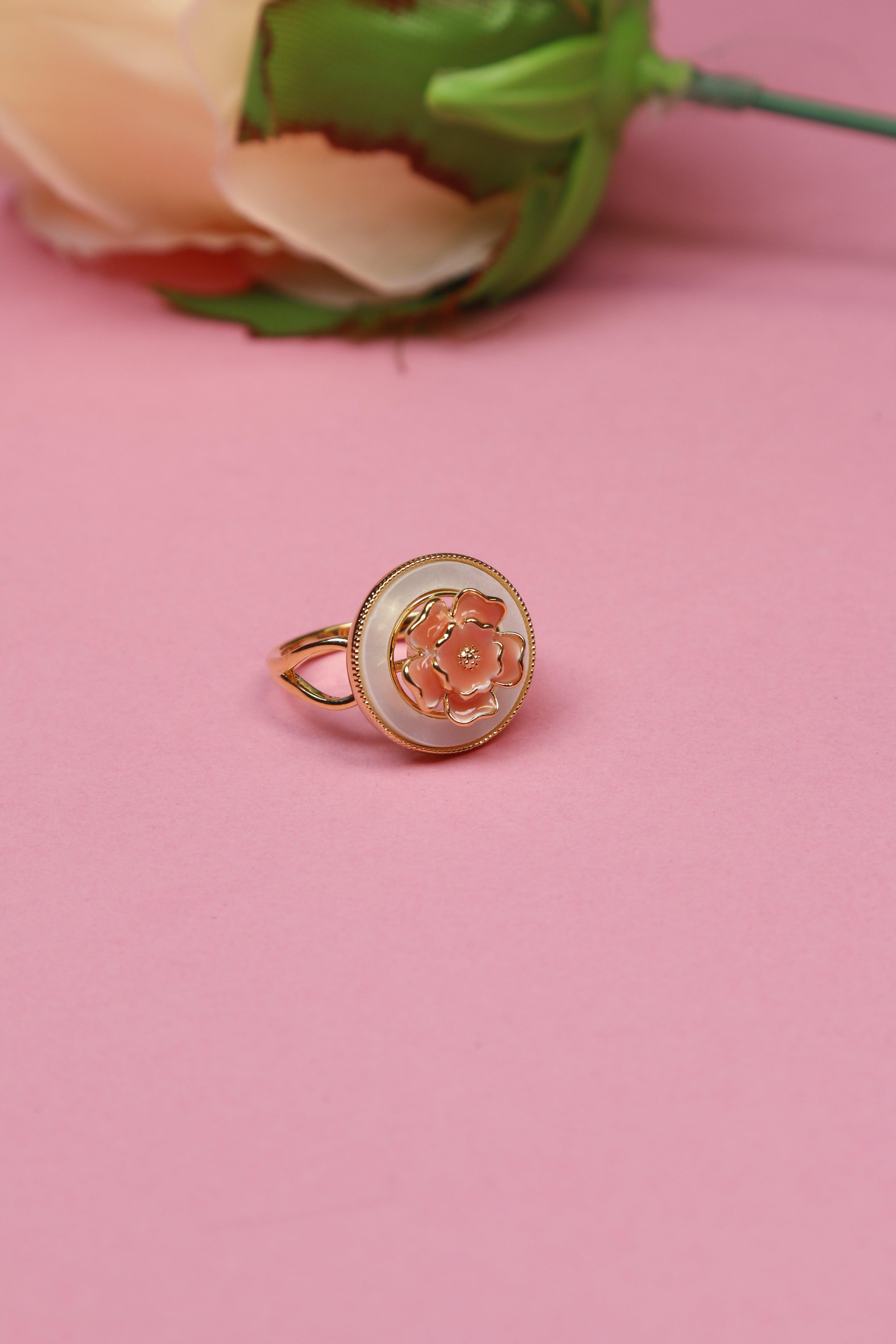 Close-up of a gold floral ring with a pink flower design set against a soft pink background. 