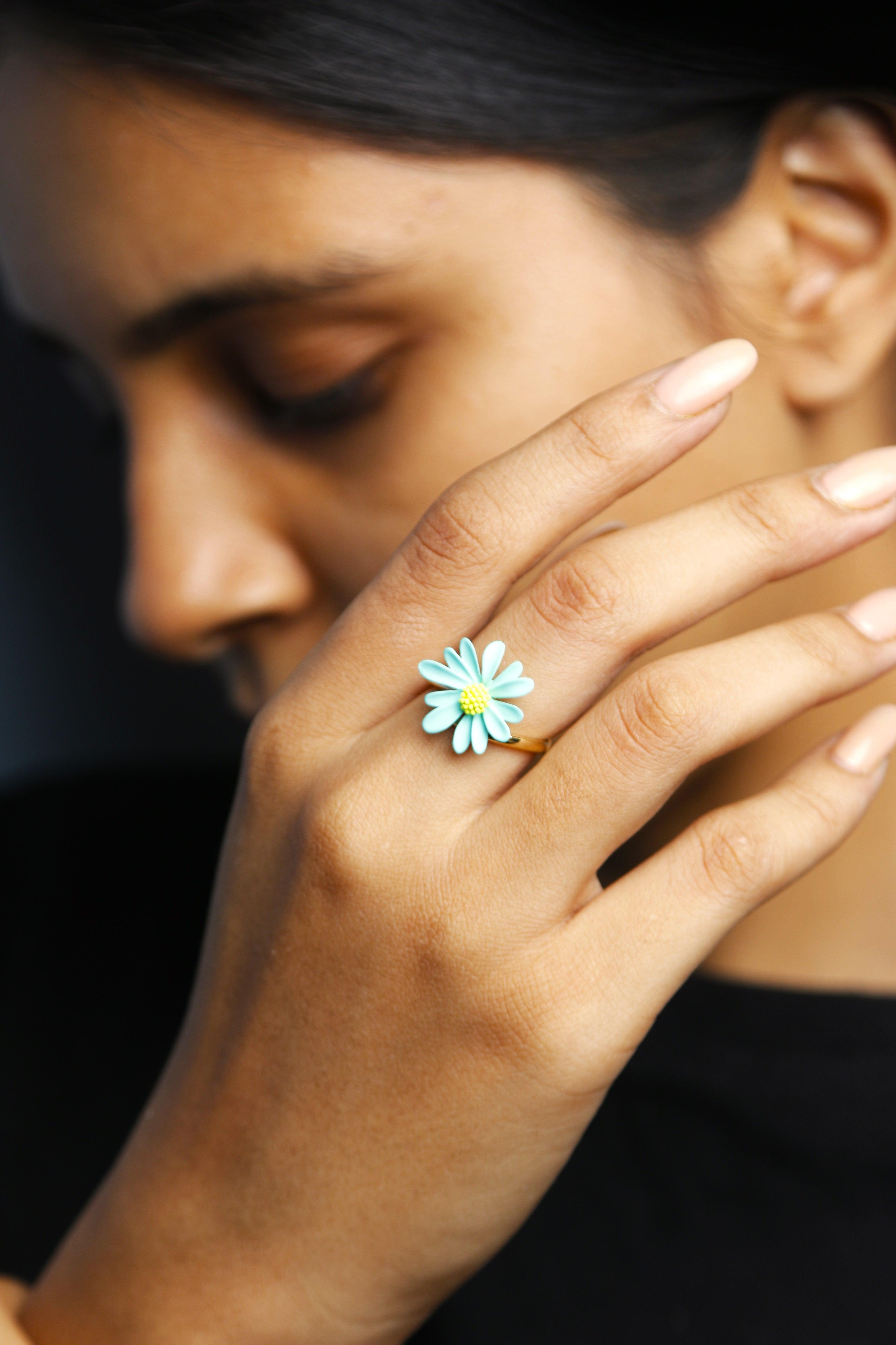 A light blue daisy flower ring with soft petals and a yellow center, placed on a finger.