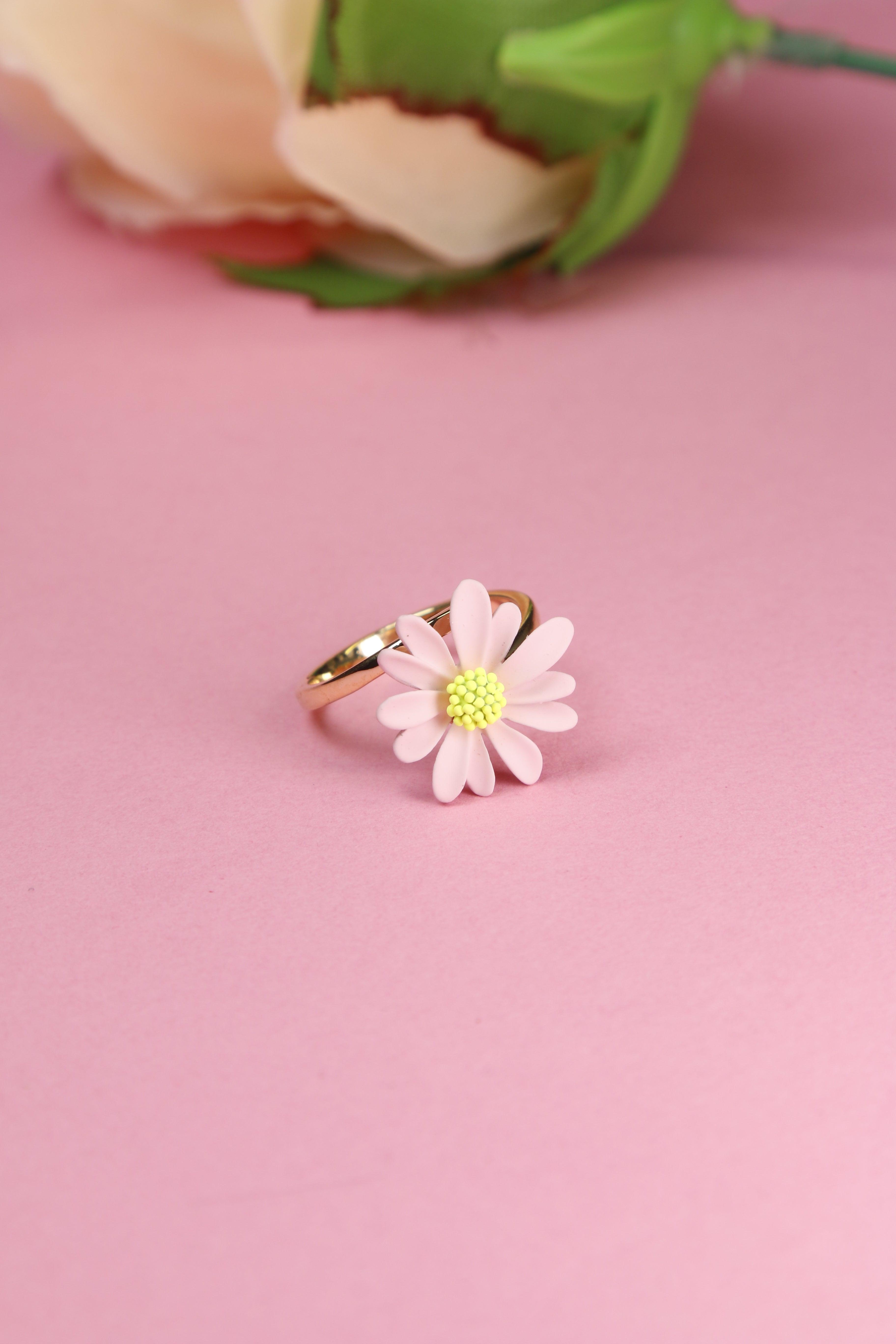 A close-up of a pink daisy ring with light pink petals and a yellow center on a gold band.